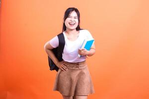 Smiling student holding a blue book and black backpack in front of an orange background photo