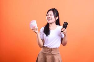 Smiling woman holding money and phone in front of orange background with space for text photo