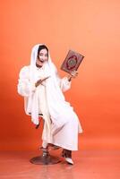 Cheerful Muslim Woman Holding Quran A Portrait of Faith and Joy, showcasing spiritual dedication with a bright smile and reverence isolated on a orange background. photo