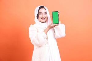 Smiling Muslim woman in hijab presents a smartphone with green screen display, happy to communicate to the user. isolated on a orange background photo