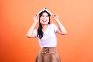 A woman with a white shirt and a brown skirt has a book on her head in front of orange background photo