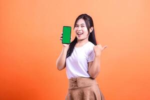 A smiling woman holds a mobile phone with a green screen and points at it with her thumb. She is wearing a white shirt and a brown skirt isolated on an orange background. photo