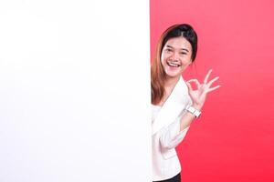 A woman in a white jacket is smiling and showing the ok okay sign while standing behind a white board, isolated on a red background photo