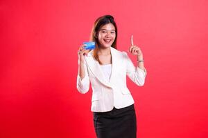 A woman in a white suit holds a credit card and points up with her index finger while smiling isolated on a red background photo
