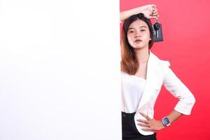 A young woman is holding car keys and posing next to a blank white panel, she's wearing a white blazer and a black pant, isolated on a red background photo