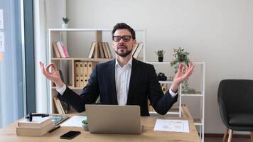 Young adult man in business attire seated at desk meditating during work pause. Scene emphasizing workplace wellness, relaxation, mindfulness. Office environment with bookshelves plants chair visible video
