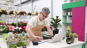 A Caucasian man in a florist shop uses a laptop at a counter, surrounded by plants and flowers. He appears to be managing inventory or sales. video