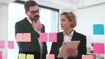 Two adult business professionals using colorful sticky notes on transparent board for teamwork planning. Male and female colleagues collaborating in modern office environment to organize video