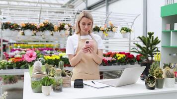 A Caucasian woman works at a florist shop, taking orders over the phone and using a laptop to manage sales while surrounded by various flowers and plants. video