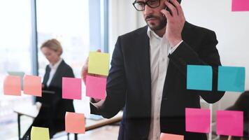 Young adult male professional in business suit uses colorful sticky notes on glass panel while talking on mobile phone in modern office environment emphasizing organization video