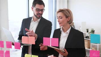 Two business professionals, a man and a woman, collaborate on project ideas using a transparent board covered in colorful sticky notes and work on digital devices in bright office setting. video