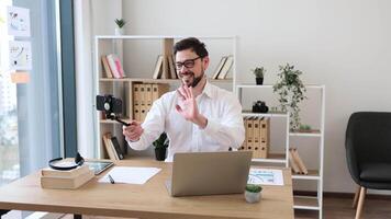 Confident man in office setting conducting call using smartphone. Young adult with glasses wearing work attire, interacting enthusiastically. Modern workplace accessories and technology video