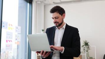 Man in early 30s wearing suit in office, holding laptop, expressing thoughtful pose, confidence. video