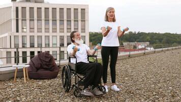 Senior man in wheelchair exercising with dumbbells assisted by woman on building rooftop. They are enjoying fitness outdoors together, focusing on health and rehabilitation in inclusive environment. video