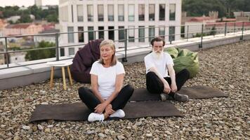 Elderly man and woman sitting in lotus pose practicing meditation on rooftop urban area. video