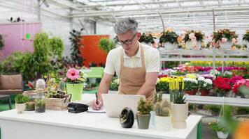A Caucasian man works at a flower shop's counter showing thumb up, surrounded by a vibrant display of plants and flowers. video