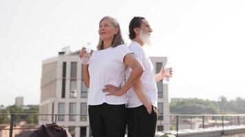 Older man and woman standing together outdoors, enjoying refreshing bottled water post workout. Rooftop setting with urban scenery creates healthy, active lifestyle atmosphere. video