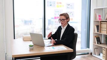 Professional middle-aged Caucasian woman in business attire communicates via call while working in modern resonant office. She is using a laptop while wearing a headset at her desk. video