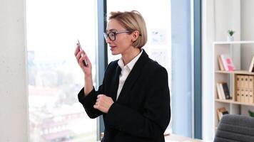 Confident woman in her 30s, dressed in formal attire, chatting on phone in stylish office setting with desk and shelves in background, conveying business success and communication video