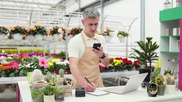A Caucasian man in an apron works at the counter of a flower shop, using a laptop and taking notes while on the phone. video