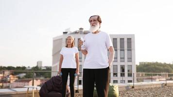 Elderly man with white beard drinking water while woman holding weights stands outdoors. Scene captured during fitness routine on rooftop showcasing importance of hydration and exercise. video