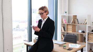 Professional businesswoman in modern office leaning on desktop browsing phone, wearing formal attire. Bright workplace with shelves, glass windows. Represents productivity, communication, multitasking video
