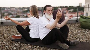 Senior man and woman seated back to back meditating in lotus pose on rooftop. Engaging in mindfulness and wellness practice. Healthy lifestyle for older adults, peaceful urban setting for relaxation. video