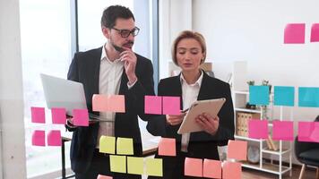 Businesswoman and businessman using colorful sticky notes on a glass board in office setting. Team collaboration involving professionals discussing project management, organizing tasks. video