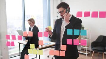 Male adult wearing suit using sticky notes on glass for organization while on phone call. Professional working environment with woman colleague in background using laptop video