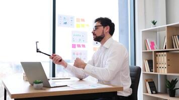 Professional businessman mid thirties with glasses seated at office desk filming call or vlog with smartphone. Bright modern workspace with shelves in background video
