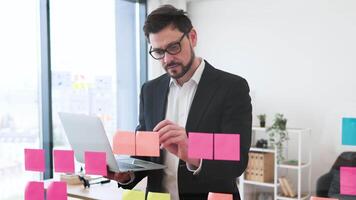 Businessman in his 30s organizing tasks on a transparent board with sticky notes, holding a laptop, working on office project planning and management, shows focus and professional work environment. video