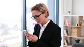 Confident middle-aged woman in formal attire standing in bright office holding smartphone smiling video