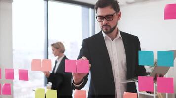 Businessman in his thirties posting sticky notes on glass board, holding laptop, conceptualizing project tasks, woman in background. Modern collaboration environment with organized, efficient process. video