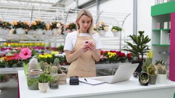 A Caucasian woman in a flower shop takes an order on the phone while writing on a notepad and using a laptop. video