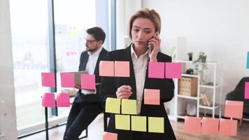 Confident adult businesswoman engaging in phone call, organizing work with colorful notes on transparent board in modern office. Male colleague visible working in background. video