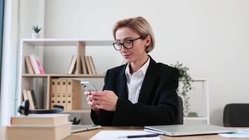 Smartly dressed businesswoman seated at desk using smartphone with happy expression. Office features organized bookshelf, papers, and plants creating professional environment. video