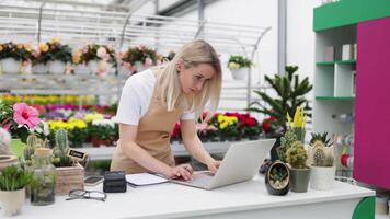 A caucasian woman works at the sales table in a flower shop, using a laptop and writing in a notebook. video