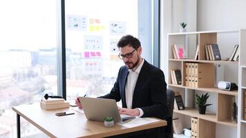Businessman wearing formal suit working at desk, planning and making notes, in modern office with bright environment and natural light. Workspace includes organized shelving and stationery. video