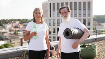 Senior man and woman smiling on rooftop, preparing for yoga or fitness activity. They carry exercise essentials and exhibit positive energy, encouraging healthy lifestyle and staying active video