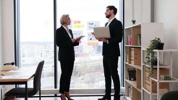 Two business professionals, one male and one female, discussing work in an office. Both are wearing formal attire, standing near a large window, with charts and diagrams visible in the background. video