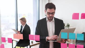 Businessman working on ideas with sticky notes on glass board, businesswoman in background. Collaborative team, modern office setting, Caucasian male and female professionals. video