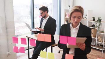 Professionals collaborating on project ideas with colorful sticky notes, utilizing a glass board for organization. Concept of teamwork and creativity in a modern office environment. video