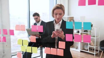 Businesswoman discussing strategy on phone near glass with sticky notes, colleague working background video