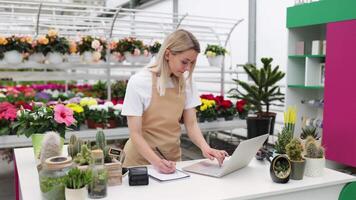 A Caucasian woman works on a laptop at a table in a flower shop surrounded by colorful plants and flowers. video