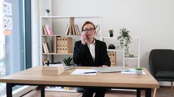 Woman enjoying phone conversation while seated at office desk in workplace demonstrating positivity. Middle-aged woman, professional attire, daytime setting with documentations, and greenery around video