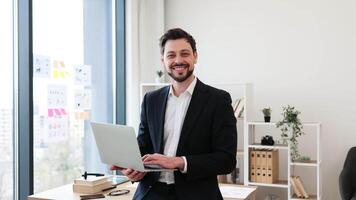 Smiling male business professional in modern office with laptop, wearing formal attire, expressing confidence and success. Young adult man, medium build, standing by desk with organized workspace video