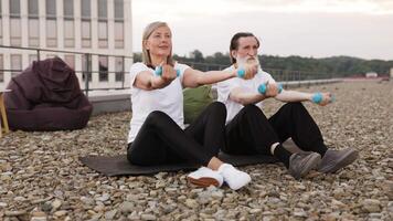 Elderly man and woman sitting on mats on rooftop doing fitness exercises with dumbbells outdoors. video