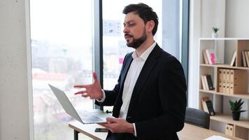 Professional adult Caucasian male in formal suit on call speaking and gesturing at laptop, in bright modern office setting. Captures positive communication, digital technology, business concept. video