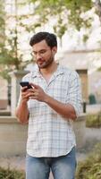 Indian young man using smartphone typing text messages in social media application online, surfing internet, relaxing, taking a break outdoors. Hispanic guy standing on urban city street. Vertical video