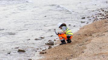 Female Engineer Using Smartphone and Laptop for Coastal Area Assessment, Environmental Scientist Conducting Beach Cleanup Survey, Worker in Safety Vest and Hard Hat Documenting Outdoor Site photo